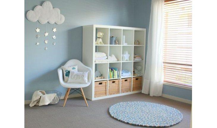 Quarto de bebê com tapete azul, estante branca e poltrona branca com itens decorativos das cores verde e azu