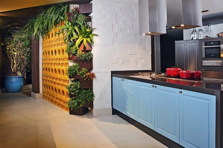 Horta vertical na parede ao lado da cozinha com plantas dentro de vasos marrons presos na parede laranja. ao lado, na cozinha, um armário azul embutido na bancada de madeira escura.