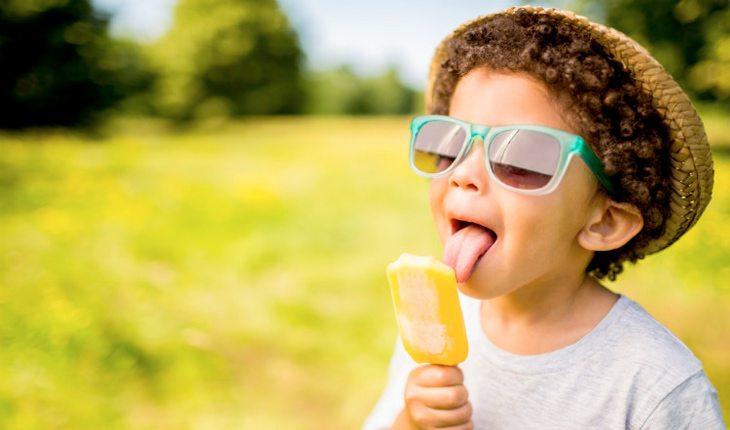 Atividades refrescantes. Na foto, um menino lambendo um picolé amarelo. Ele está usando um óculos de sol e um chapéu