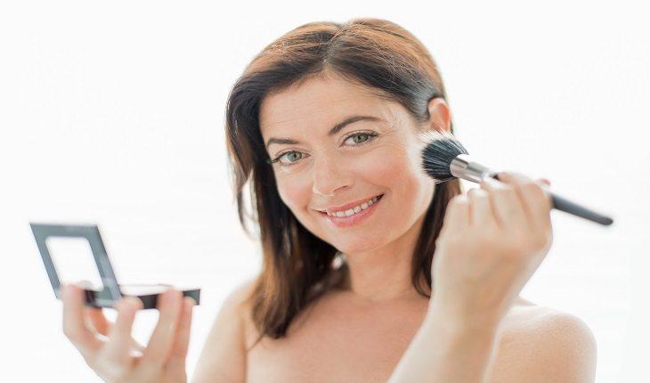 mulher sorrindo passando blush com um pincel de maquiagem e segurando um espelho de mão