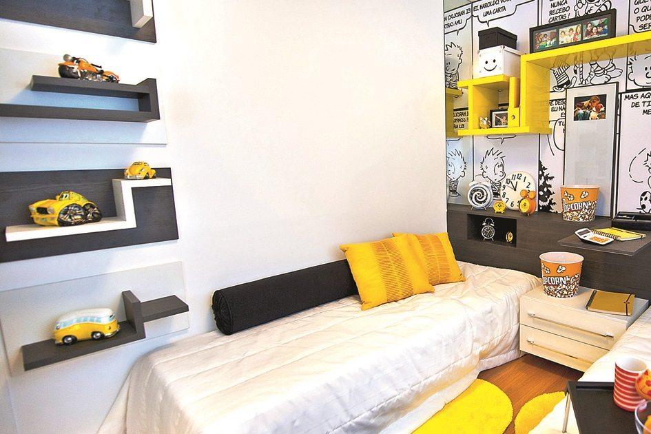 Cama branca com almofadas preta e amarelas, parede la direita toda adesivada com cenas de quadrinhos e na parede branca, prateleiras pretas com artigos decorativos.