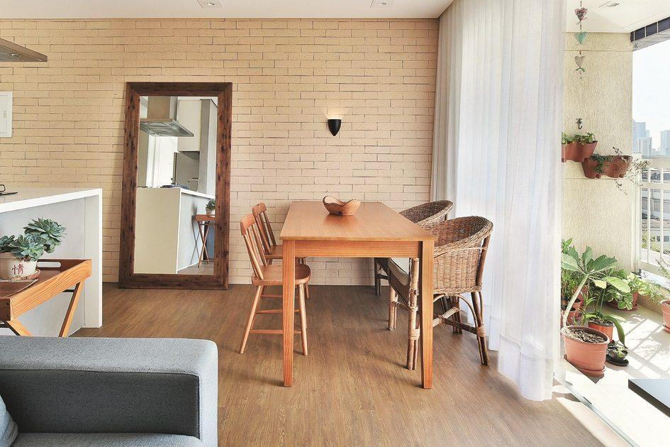 Ambientes unidos com uma mesa de madeira de 4 lugares ao lado do sofá cinza. Do lado esquerdo da mesa, há um espelho grande no chão com moldura de madeira.