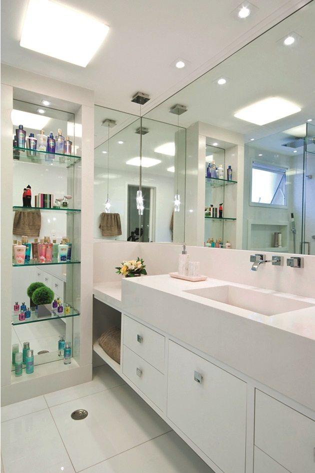 Banheiro todo branco, com grande espelho dando sensação de amplitude. Do lado esquerdo, prateleiras de vidro que organizam os cosméticos transformando os banheiros.