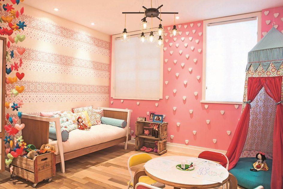 Uma parede listrada em branca e rosa e outra rosa com textura de corações brancos. Ao lado da cama à um criado mudo feito com um caixote para dar diversão para as crianças.