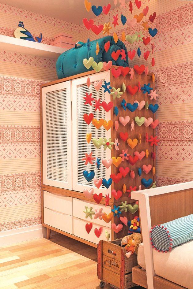 Armário de madeira com portas brancas no canto do quarto infantil feminino, com um enfeite pendurado no teto com vários corações coloridos criando um ar de diversão para as crianças.