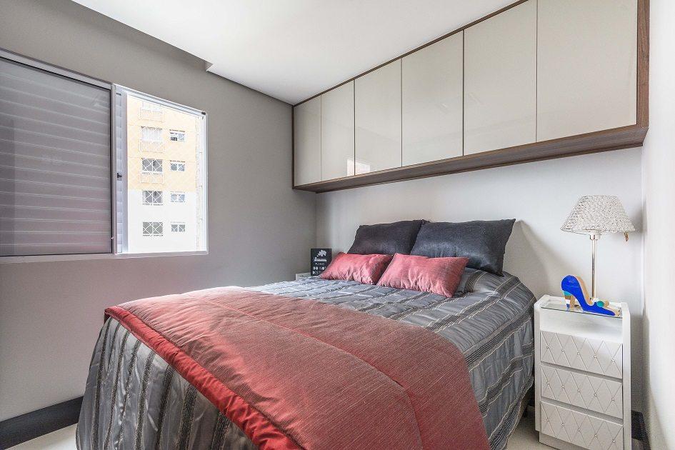 Um dos quartos de casais com uma cama de casal com edredom e almofada cinza e vermelho. Acima da cama há um armário em marcenaria planejada em tons claros.