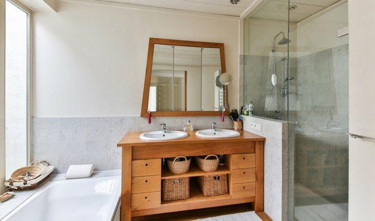 Foto panorâmica de um banheiro pequeno com o revestimento em cimento queimado e pastilhas, com um box feito de vidro, uma pia branca em porcelanato e um espelho redondo com moldura de madeira.