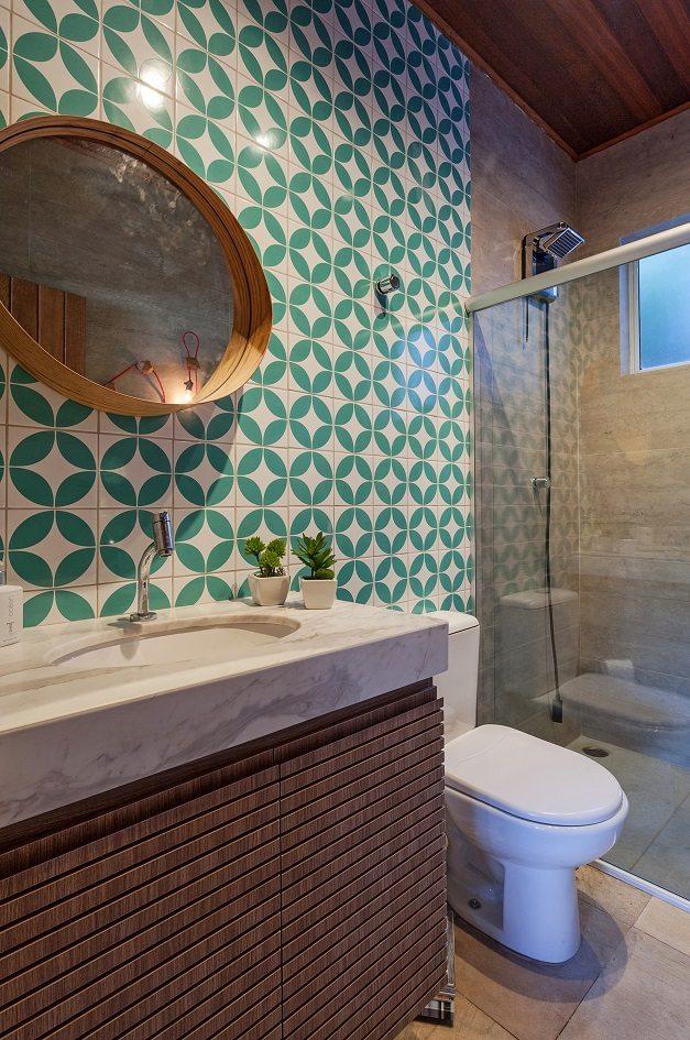 Foto vertical de um banheiro com o revestimento da parede direita em cimento queimado e da esquerda com pastilhas em formatos geométrico em branco e verde.
