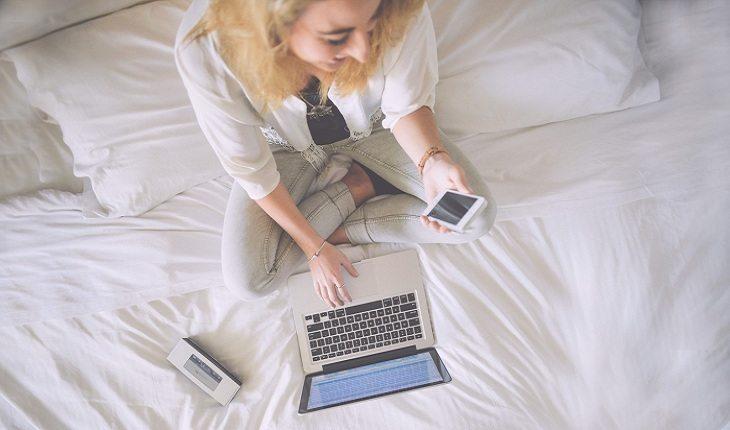 foto de uma mulher na cama, mexendo no celular e perto do computador
