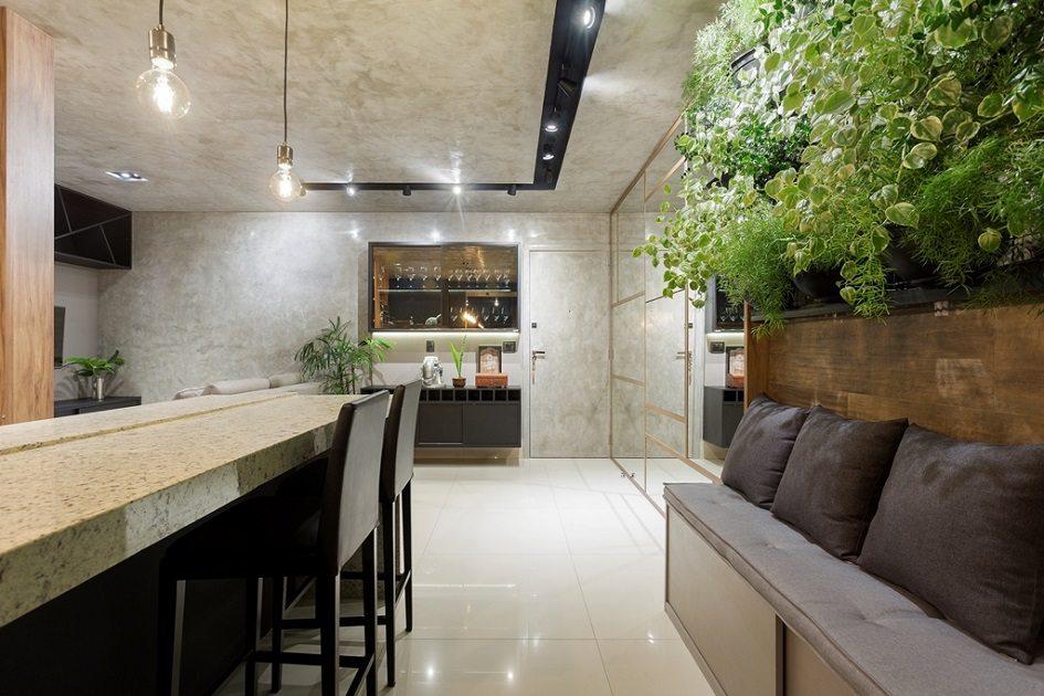 Cozinha com um jardim vertical do lado direito e embaixo um sofá feito em marcenaria multiúso. Do lado esquerdo há uma bancada com dois bancos altos.