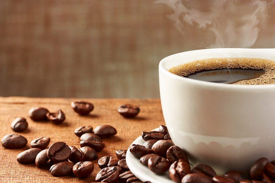 Xícara com café branca em uma mesa de madeira e fundo marrom com grãos de café espalhados pela mesa e no pires que podem prejudicar o detox emagrecedor.