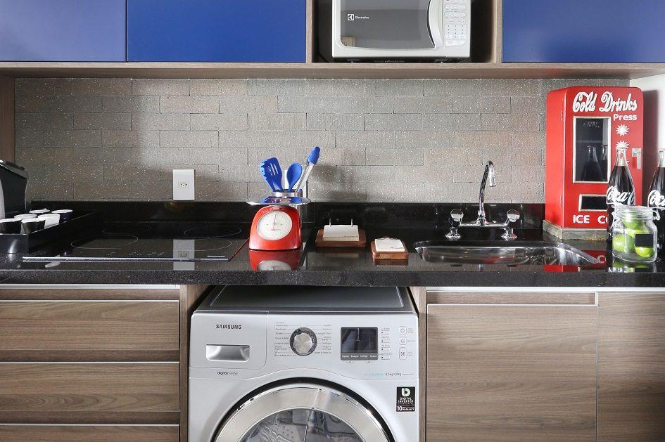 Como organizar a lavanderia com um armário planejado na cor azul, com paredes feitas de tijolinhos e a máquina de lavar abaixo da bancada da cozinha.