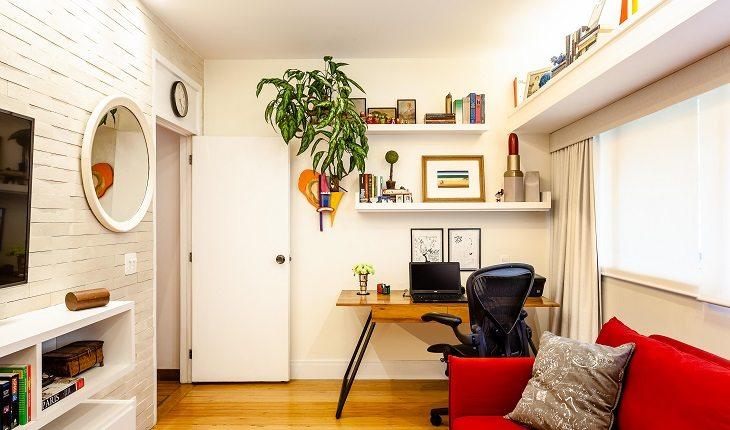 Mesa para escritório ao lado da janela com prateleiras, vaso de planta suspensa, sofá cama.