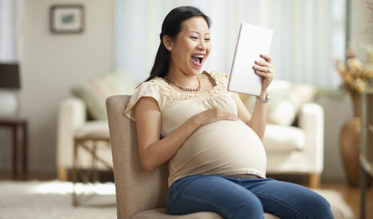 Na imagem, a mulher está segurando a barriga grávida sorrindo com um tablet na mão. Idade para engravidar.