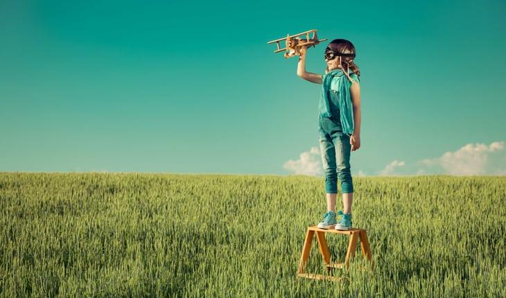 Menina em cima de banco em um campo gramado segurando um avião e com óculos de aviadora