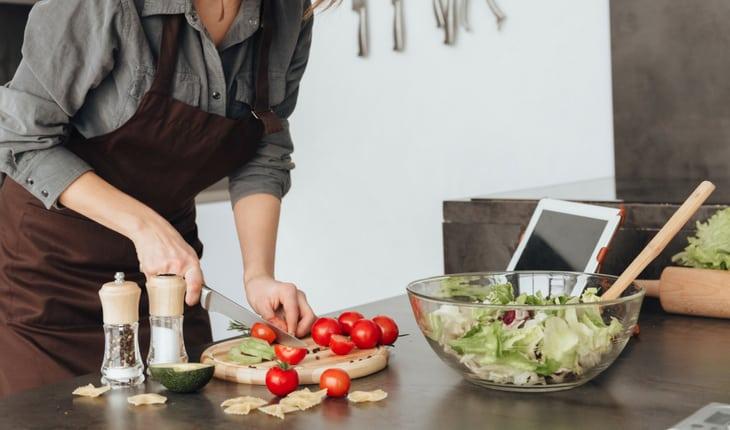 Mulher de avental picando tomates em tábua com bowl de saladas com utensílios de cozinha ao lado