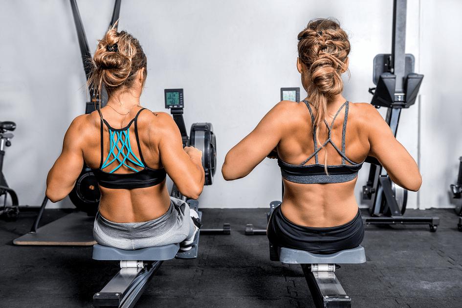 Duas mulheres de costas e sentadas em uma máquina de remar na academia para ilustrar o tema exercícios para costas e ombros