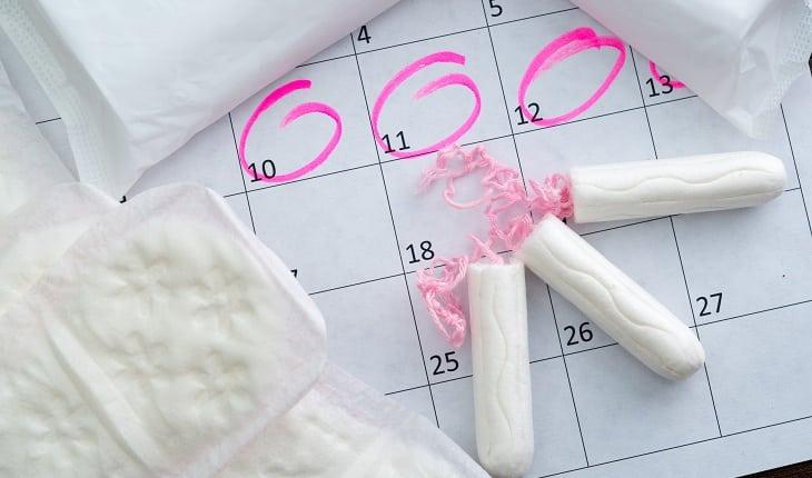 Na imagem, diversos tipos de absorvente está disposto em cima de um calendário, com datas marcadas em rosa. Cólica menstrual intensa.