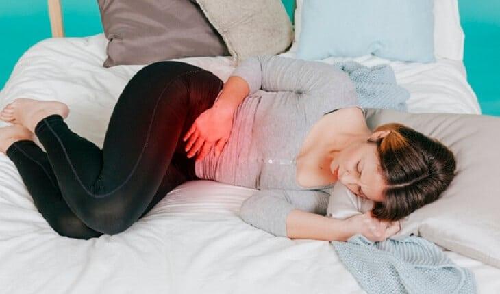 Na imagem, a mulher está na cama deitada com dores na barriga. Cólica menstrual intensa.