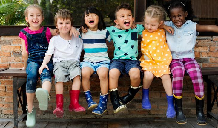 Crianças loiras, japonesas e negras sentadas em banco abraçadas e dando risada