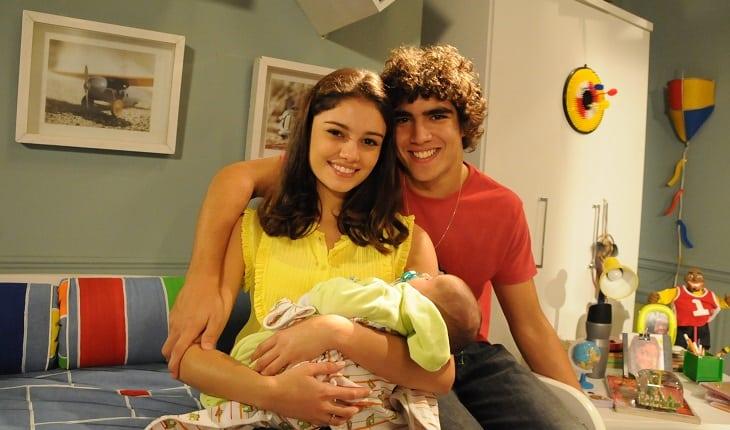 Casais de Malhação: Bruno e Angelina. Foto posada em que a personagem segura um bebê e o namorado está ao seu lado com o braço envolta dela.