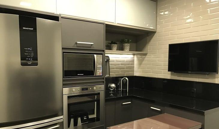 Foto de uma cozinha com armários escuros, eletrodomésticos em inox e mesa na cor berinjela. Há uma TV na parede lateral da mesa