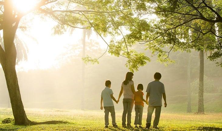 A foto mostra um pai, uma mãe e seus dois filhos de mãos dadas sob uma árvore em um parque