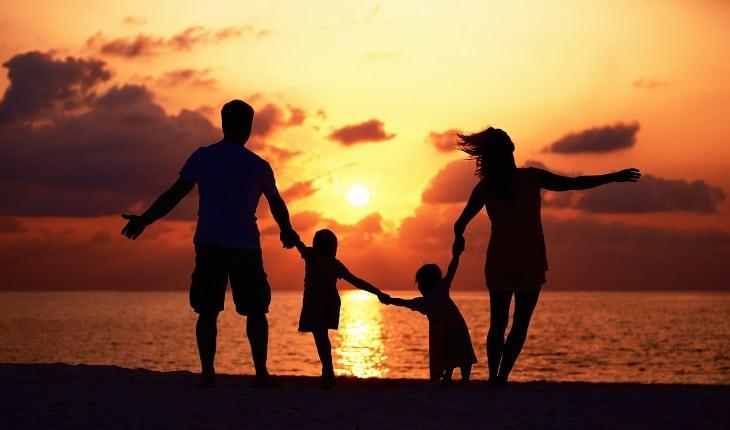 A foto mostra um pai e uma mãe de mãos dadas com seus filhos em frente ao mar