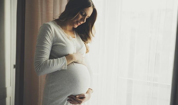A foto mostra uma mulher grávida olhando para sua barriga e sorrindo