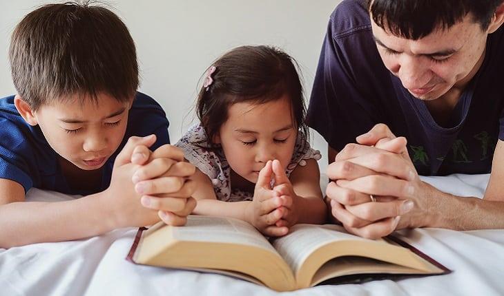 A foto mostra um pai lendo a Bíblia e orando com seus dois filhos pequenos