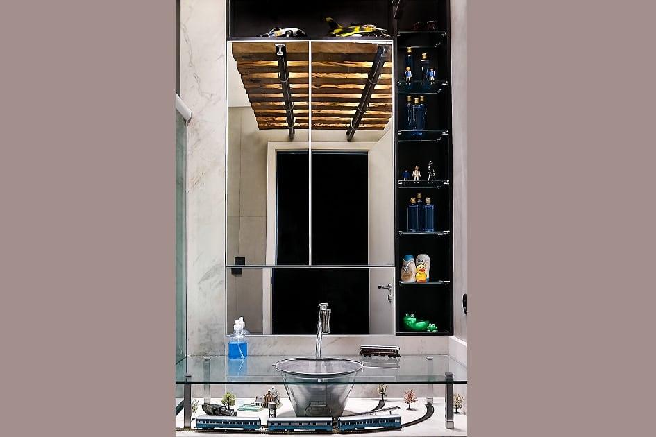 Foto da pia do banheiro, na qual aparece a bancada em vidro, a pia que é um balde prateado reciclado e o espelho, que reflete os trilho do trem no teto