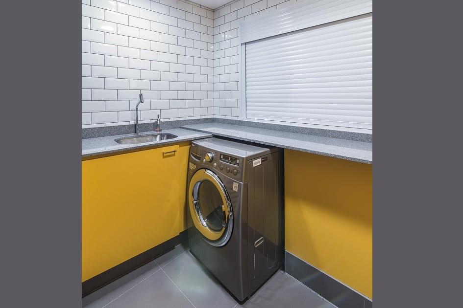 Foto da lavanderia do banheiro, na qual a revestimento metrô white nas paredes, a bancada é cinza, os armários amarelos e a máquina de lavar roupa é em inox