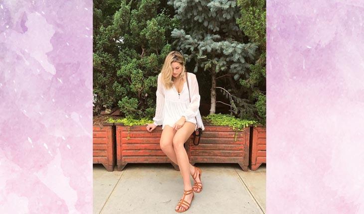 Sasha com uma blusa branca e com uma sandália marrom sentada no jardim